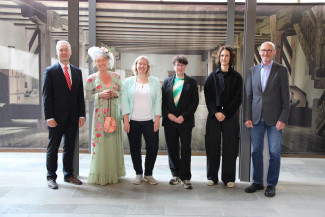 Gruppenbild mit sechs Männer und Frauen mit Kunstfigur Henriette Huch in grünem Kleid mit Hut
